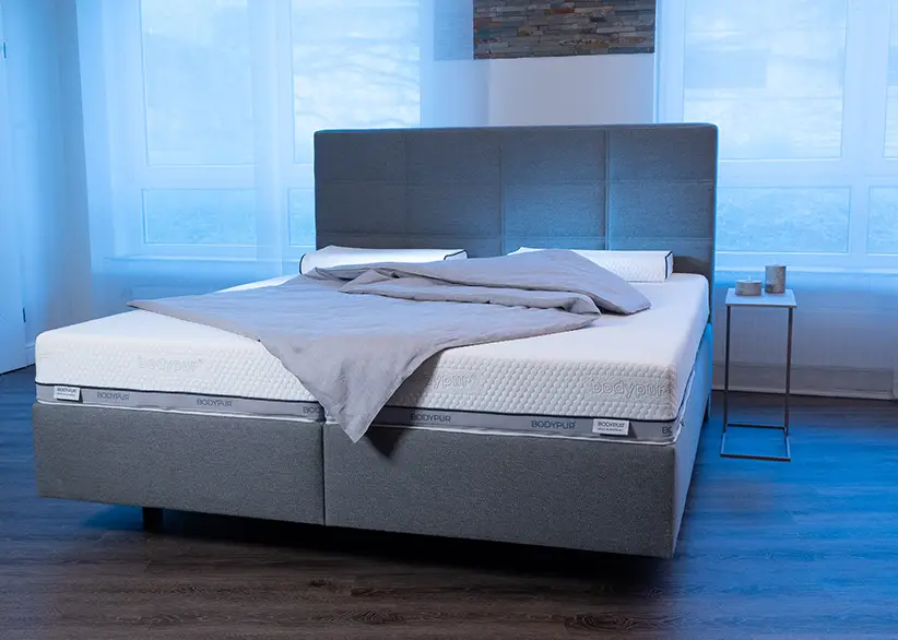 Eine Bodypur-Matratze und Kopfkissen arrangiert auf einem Bett mit grauem Kopfteil und blauen LED-Leuchten im Hintergrund, was eine beruhigende Schlafumgebung suggeriert.